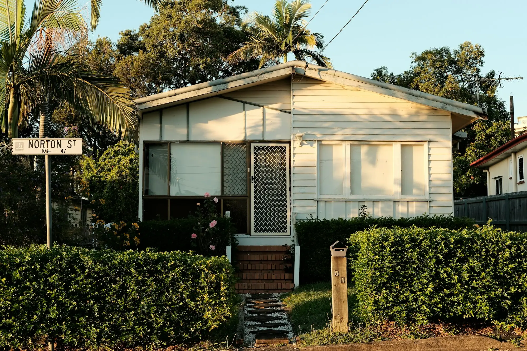 Post-war fibro cottage behind a hedge on a suburban street with a corner street sign